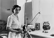 A woman in a short-sleeved dress, using a camera and tripod to photograph a Greek vase