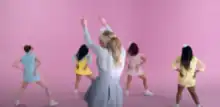 Four women twerking and one standing still in front of pastel-colored pink backdrop.