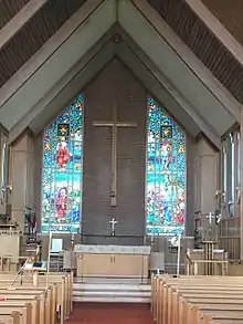 The interior of a building, with a vaulted ceiling supported by wood beams. The far end shows the back wall, which has a large wooden cross hung on a wall between two stained glass windows. An altar is visible before the wall, with a central path leading from it to the foreground, adjacent to which are rows of pews.