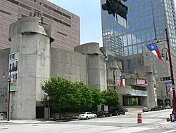 Concrete building in two sections, each with a five-sided tower on the ends. One is a curved and windowless. The other looks like a theater entrance with rounded marquee shapes and glassed entrance.