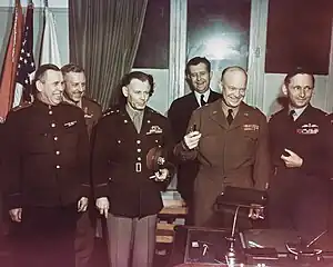 Group shot of six smiling men (and one barely visible woman), each in a different military uniform, standing behind a writing desk. Eisenhower is holding three fountain pens. In the background are flags, including the US flag.