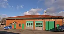 A large, sunlit, single-storey orange brick building with a roof covered with red tiles. On the left are two small green doors, in the centre are three large, green, shutter doors. Atop the roof is an aerial.