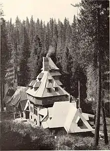 A monochrome photograph of a 75-foot-tall building with castle-like features, seen from a nearby higher elevation, surrounded by tall pine trees.