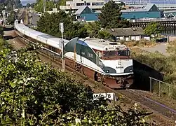 A passenger train with white, dark brown, and dark green stripes. The green stripe curves up along the side of the diesel locomotive.