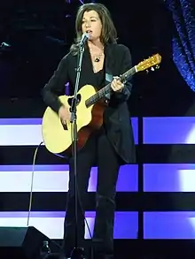 Standing, Grant sings into a microphone while playing an acoustic guitar