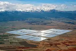 Concentrated solar power parabolic troughs in the distance arranged in rectangles shining on a flat plain with snowy mountains in the background