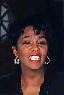 A woman sings while she is recharged on a piano. She wears a long black ensemble and diamond earrings.