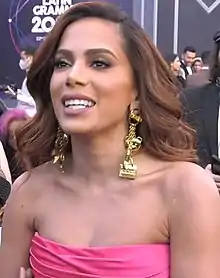 A woman with large dangling gold earrings and a strapless pink dress smiles at an awards ceremony while people loiter behind her.