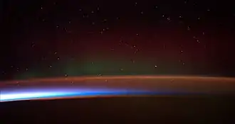 Earth's night-side upper atmosphere appearing from the bottom as bands of afterglow illuminating the troposphere in orange with silhouettes of clouds, and the stratosphere in white and blue. Next the mesosphere (pink area) extends to the orange and faintly green line of the lowest airglow, at about one hundred kilometers at the edge of space and the lower edge of the thermosphere (invisible). Continuing with green and red bands of aurorae streching over several hundred kilometers.