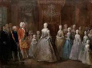 A throne room with a middle-aged lady in a silver dress standing in the middle, facin a man in a gilded red coat. Between them is a little girl in a light blue dress and a boy. Behind the lady stand five young girls, descending in height towards the edge of the canvas. Many courtiers are standing in the bacgkround.