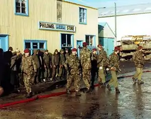 A group of armed soldiers in camouflage uniform with red berets guard a line of enemy soldiers, who stand in front of a large wooden building, which is painted yellow. The road is wet, and the sky is blue.