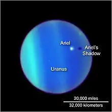 the planet Uranus is seen through the Hubble telescope, its atmosphere defined by bands of electric blue and green. Ariel appears as a white dot floating above it, casting a dark shadow below