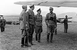 an Italian officer and three German officers in uniform standing beneath the wing of an aircraft on a grassed airfield