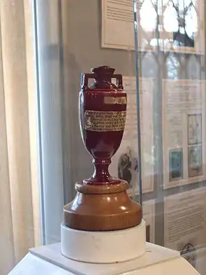 A terracotta urn with two brass plaques on it sits on a wooden base. The urn is on display, with the glass surround and other exhibits visible in the background.