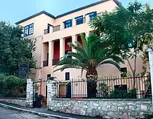 Photograph of a sand-coloured house with a fence outside and palm trees growing in the garden.