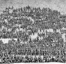 Black and white photo of a large number of men wearing military uniform seated on stone steps