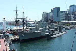 A destroyer and a submarine tied up alongside a wharf. The destroyer is next to the wharf, with the submarine docked alongside the destroyer. A sailing ship is moored on the other side of the wharf. Several small boats are underway around and behind these three vessels.