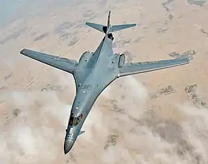 Top view of B-1B in-flight with white clouds scattered underneath. Its wings are swept fully forward.