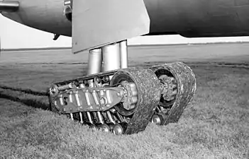 The experimental tracked landing gear unit of an XB-36, consisting of 2 flexible tracks supported by wheels, is pictured stopped in short grass with ruts trailing behind it.