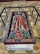 A stone tomb, with the effigy of a robed man on top, with a coat of arms at his feet displaying a red panther on a blue-and-gold striped background.