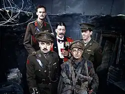 The main members of the cast of Blackadder Goes Forth are seated in the production's World War I trench set. Seated in the centre is Stephen Fry as General Melchett, incongrously wearing red officer's mess dress and holding a sherry. Tim McInnerny as Captain Darling and Hugh Laurie as Lieutenant George stand beside him in green officers' uniforms. Most prominent in the picture is Rowan Atkinson as Captain Blackadder, wearing his captain's uniform. To his right is Tony Robinson as Baldrick, wearing a moth-eaten private's uniform, his cap angled off-centre. George is the only one smiling.