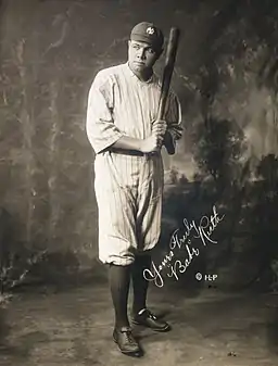 A man in full baseball attire wears a pinstriped jersey and a baseball cap. Looking to the left of the camera, he is holding a baseball bat upward.