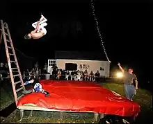 A backyard wrestler falling towards a cloth covered table as his opponent rolls out of the way of the move.