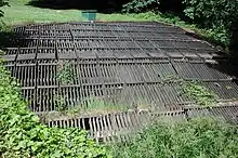 A horizontal wooden rack roughly 30 feet (9.1 m) wide and 30 feet (9.1 m) long lies in flat, grassy area.