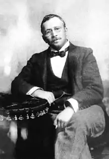 Black and white portrait of a seated man. He has a short beard, is wearing glasses and is dress formally.