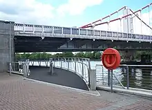 Start of a footbridge that curves under one end of a suspension bridge, along the bank of a wide river.