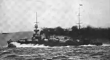 A large black ship, partially obscured by thick smoke from its funnels, creates a large bow wave as it plows through the water at high speed.