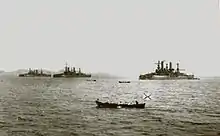 A black and white shot of three large ships near a hill-like background, with several small skiffs in the foreground