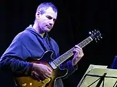 A man in a sweatshirt holding a guitar and looking down at sheet music on a stand