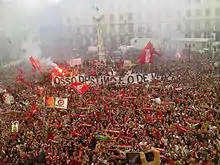 A crowd of Benfica supporters fills the Lisbon City Square. Scarves, flags, banners, and flares can be seen from the City Hall's central balcony.