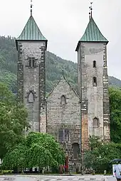 Stone  Romanesque church with two towers and a lower, pitched roof entrance hall between them. The towers have slightly curved bronze roofs.