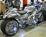 A motorcycle with fully enclosed bodywork painted with a complex black, gray and white pattern, and crome plating on many parts of the bike, and an extended length rear swingarm and an extraordinarily wide rear wheel, indoors with special chrome wheels on display in the background.