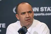 A man in a white shirt (Penn State coach Bill O'Brien) sits in front of a blue background with the words "Penn State" partly visible. He is addressing the media in front of a microphone emblazoned with the Penn State logo.