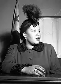 African-American woman, in her late thirties, sitting in a courtroom, looking worryingly to the right. She is wearing a feather hat, a dark coat, and a large ring on her right hand resting upon the witness stand.