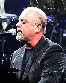 A man with a goatee and his eyes closed behind a microphone on a piano, wearing a black shirt and striped jacket.