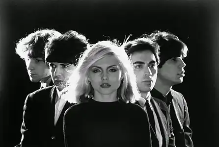 A black and white photo of Blondie posing in a triangular position over a black background.