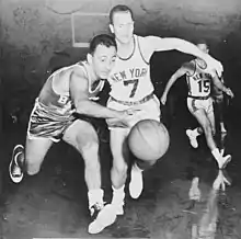 Two basketball players are going after a basketball, while another player are watching from behind.