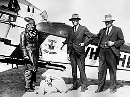 Three men, two in suits and hats, the other in flying gear, standing in front of biplane bearing the legend "Royal Air Mail"