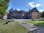 Photo a long, two-storeyed house with exposed wooden beams, many gables and small-paned windows. In the foreground is a lawn and trees are on the left.