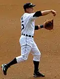 Baseball player in white uniform in a baseball throw followthrough position.  His arm is forward with his hand down and his left hand is gloved. His left foot is planted and his right foot is in the air behind him.