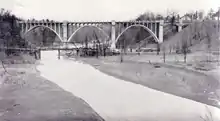 "A black and white photograph of two bridges crossing a valley. The bridge in the background towers over the valley, concrete arches rising high from the ground. The bridge in the foreground is minuscule in comparison, a tiny iron bridge over the creek running through the valley."