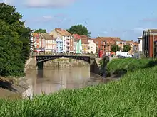 Metal bridge over the river. In the background are coloured houses and several trees.