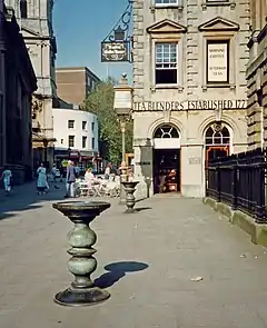 Two ornate metal pillars with large dishes on top in a paved street, with an eighteenth-century stone building behind, upon which can be seen the words "Tea Blenders Estabklishec 177-". People sitting at café-style tables outside. On the right are iron railings.