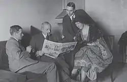 Black-and-white photograph of three men in suits and one woman seated in a room and looking at an open newspaper