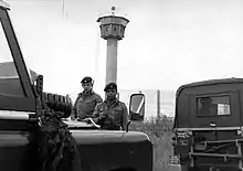 Two British soldiers carrying rifles standing behind a pair of Land-Rover vehicles, one of which has a "British Frontier Service" plate. Behind them is a high mesh fence, behind which is a tall watchtower with an octagonal cabin at its top.