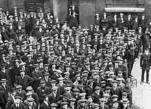 A large throng of people congregate, surrounded by police officers. Approximately half wear peaked caps; the rest wear boaters. Some smile, the rest look pensive.